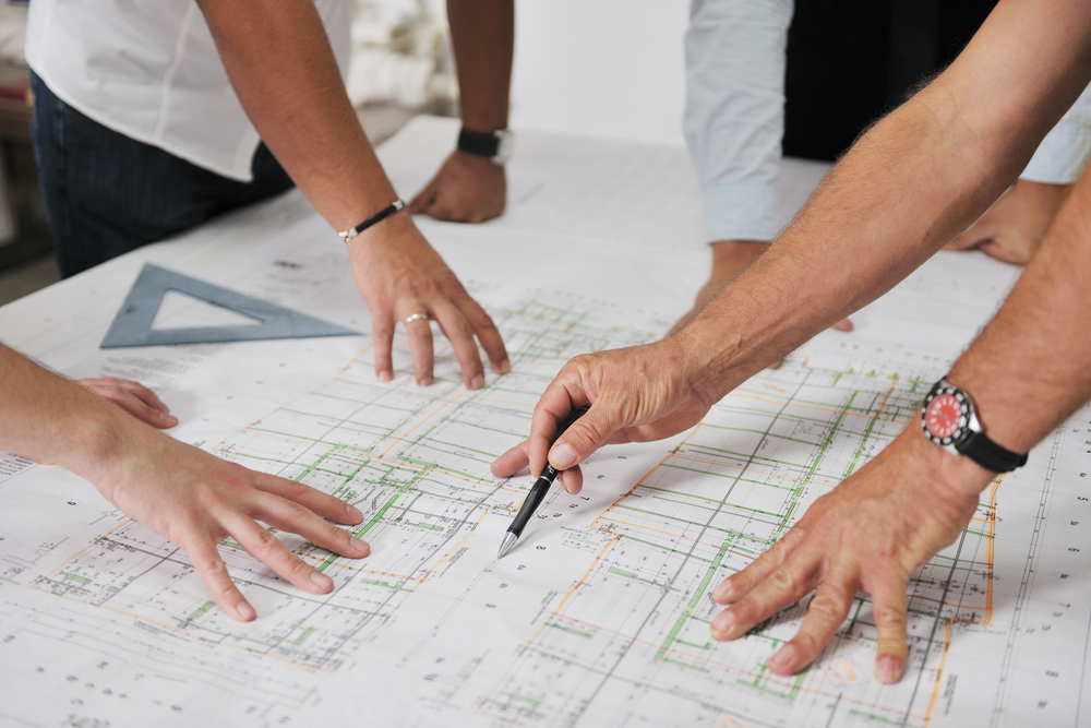 A group of construction estimators stand over an architects drawings. We only see their hands. They're measuring using a pen and scale rule