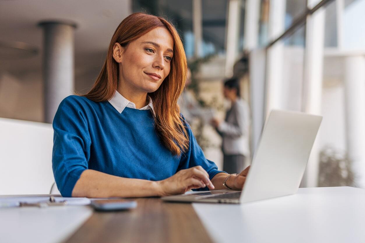 Young woman with red hair, wearing blue top sits at desk with open laptop